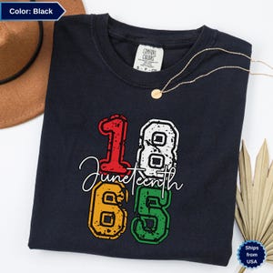 Op de afbeelding: Een zwart t-shirt met een verweerd grafisch ontwerp met het rode cijfer 1, het witte cijfer 8, het gele cijfer 6 en het groene cijfer 5. Het woord Juneteenth staat in witte cursieve letters over het midden van de cijfers.