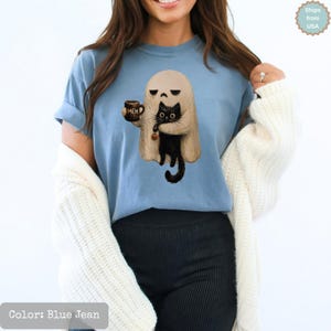 May include: A blue jean colored t-shirt featuring a graphic of a beige ghost holding a brown mug with the text MEH and a black cat. The ghost has a neutral expression and the cat has large eyes. The shirt has short sleeves and a crew neckline.