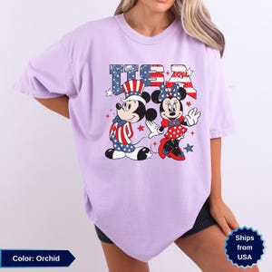 May include: Orchid-colored t-shirt featuring Mickey and Minnie Mouse in patriotic attire. The word "USA" is printed above the characters in a red, white, and blue design. The shirt is a comfortable, oversized fit.
