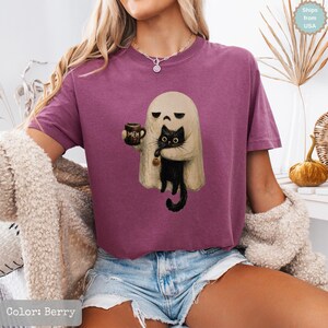 May include: A berry-colored short-sleeve t-shirt featuring a graphic of a beige ghost holding a brown mug with the text MEH and a black cat. The ghost has a neutral expression and the cat has wide eyes. The shirt has a crew neckline.