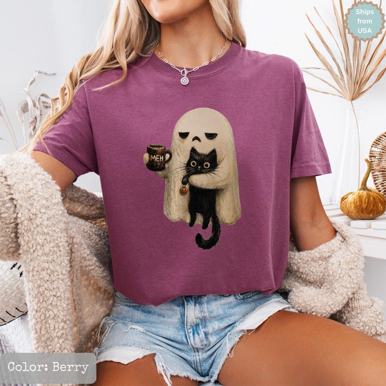 May include: A berry-colored short-sleeve t-shirt featuring a graphic of a beige ghost holding a brown mug with the text MEH and a black cat. The ghost has a neutral expression and the cat has wide eyes. The shirt has a crew neckline.