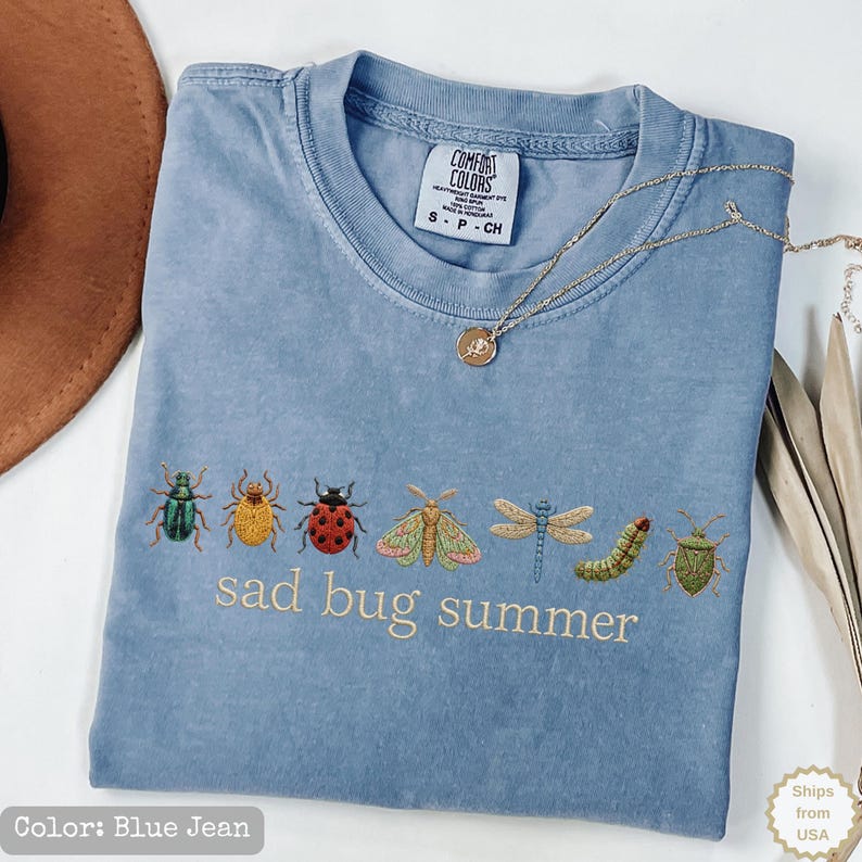 May include: A blue jean colored t-shirt featuring an embroidered row of various insects including a beetle, ladybug, moth, dragonfly, and caterpillar, with the text "sad bug summer" stitched in light thread below the insect graphic.