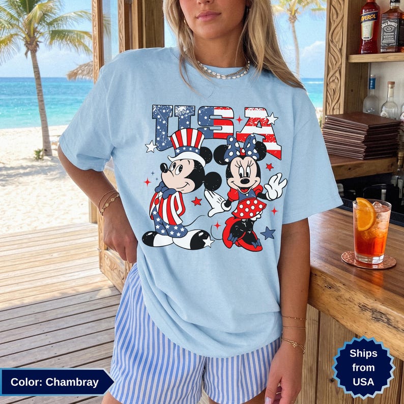 May include: Light blue chambray t-shirt featuring Mickey and Minnie Mouse in patriotic attire with the word "USA" in red, white, and blue. The design includes stars and stripes, perfect for celebrating the Fourth of July.