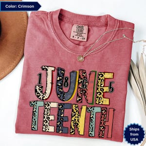Op de afbeelding: Kleurrijk T-shirt in de kleur karmozijnrood met de tekst "June Teenth" in een kleurrijk luipaardprint ontwerp. Het shirt heeft een ronde hals en korte mouwen. Een zilveren ketting met hanger ligt over het shirt.