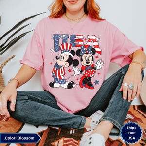 May include: A light pink t-shirt featuring Mickey and Minnie Mouse in patriotic attire with the text "USA". Mickey Mouse wears a hat and scarf in red, white, and blue. Minnie Mouse wears a red and white polka dot dress.