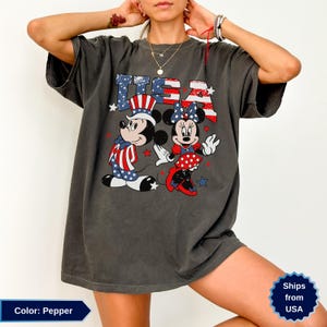 May include: Pepper-colored t-shirt featuring Mickey and Minnie Mouse in patriotic attire. The graphic includes the text "USA" in red, white, and blue, with stars. The design showcases Mickey in a Uncle Sam hat and Minnie in a polka dot dress.