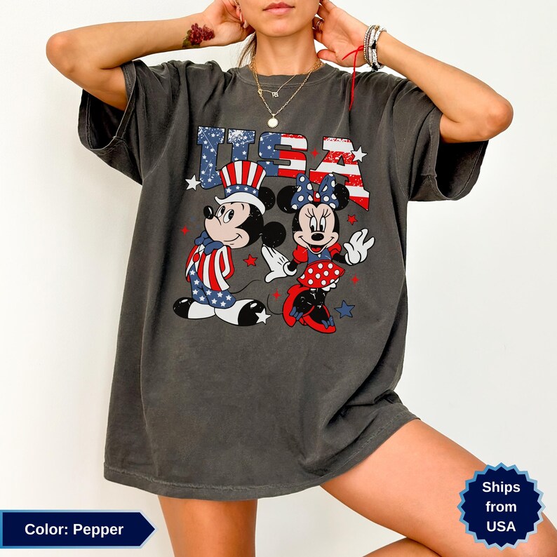 May include: Pepper-colored t-shirt featuring Mickey and Minnie Mouse in patriotic attire. The graphic includes the text "USA" in red, white, and blue, with stars. The design showcases Mickey in a Uncle Sam hat and Minnie in a polka dot dress.