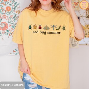 May include: A mustard yellow short-sleeve t-shirt featuring a horizontal row of various colorful insect illustrations, including a beetle, ladybug, moth, dragonfly, and caterpillar, with the black text "sad bug summer" printed below.