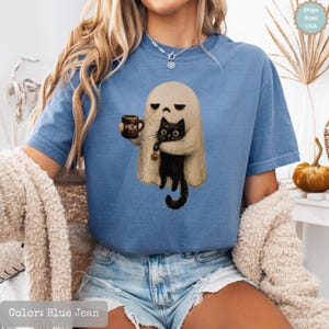 May include: A blue jean colored t-shirt featuring a graphic of a beige ghost holding a brown mug with the text MEH and a black cat. The cat is holding a small orange pumpkin. The shirt has short sleeves and a crew neckline, worn with denim shorts.