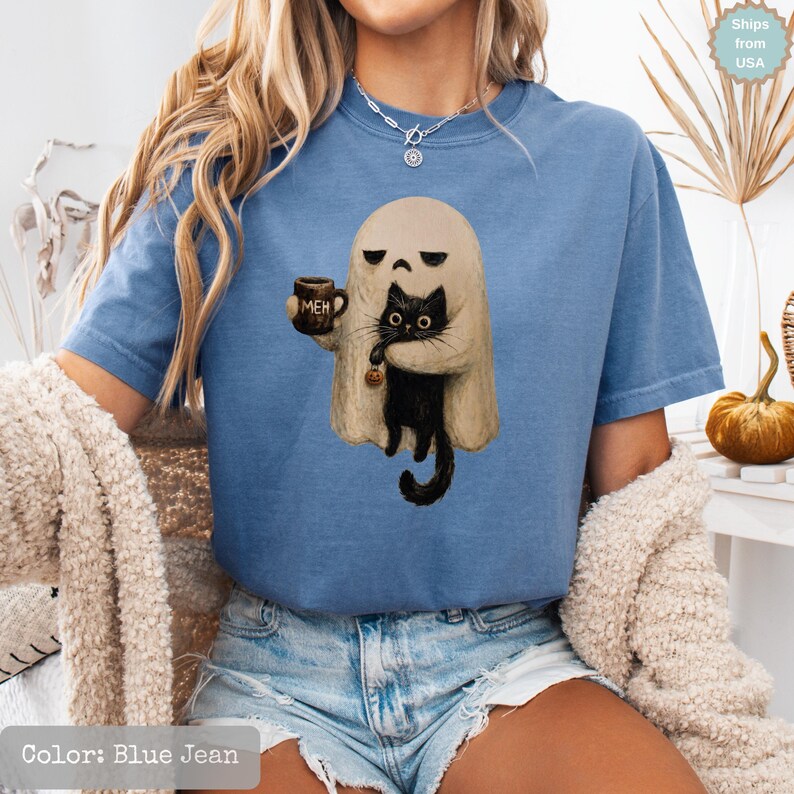 May include: A blue jean colored t-shirt featuring a graphic of a beige ghost holding a brown mug with the text MEH and a black cat. The cat is holding a small orange pumpkin. The shirt has short sleeves and a crew neckline, worn with denim shorts.