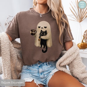 May include: An espresso-colored short-sleeve t-shirt featuring a graphic of a beige ghost holding a black cat and a mug with the text MEH printed on it. The ghost has a sad expression, and the cat is holding a small orange pumpkin charm.