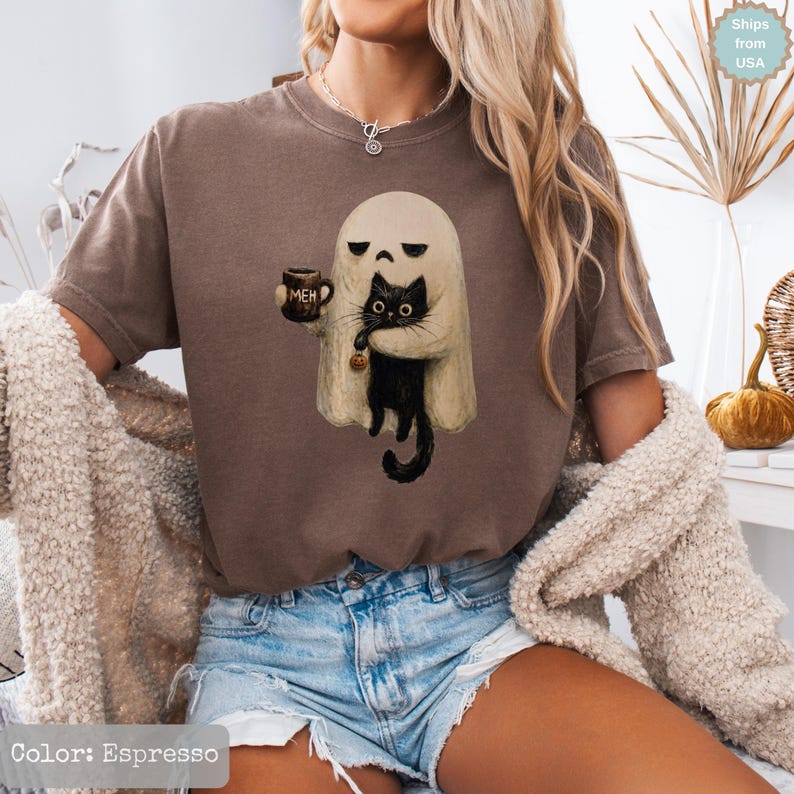 May include: An espresso-colored short-sleeve t-shirt featuring a graphic of a beige ghost holding a black cat and a mug with the text MEH printed on it. The ghost has a sad expression, and the cat is holding a small orange pumpkin charm.