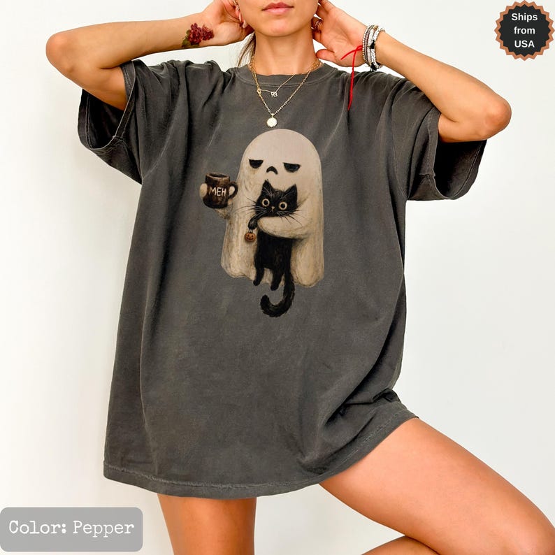 May include: A pepper-colored t-shirt featuring a graphic of a white ghost holding a black cat and a mug with the text MEH printed on it. The ghost has a neutral expression, and the cat has large eyes. The shirt has short sleeves and a crew neck.