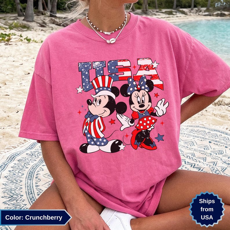 May include: Crunchberry-colored t-shirt featuring Mickey and Minnie Mouse in patriotic attire with the word "USA" in red, white, and blue. The design includes stars and stripes, perfect for celebrating the Fourth of July.