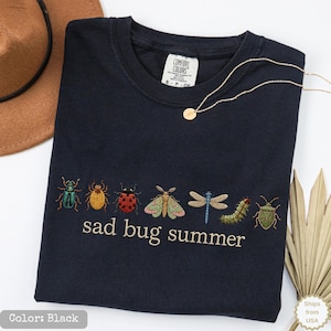 May include: A black short-sleeved t-shirt featuring an embroidered row of colorful insects including a beetle, ladybug, moth, dragonfly, and caterpillar, with the text "sad bug summer" embroidered in white thread below the insect graphic.