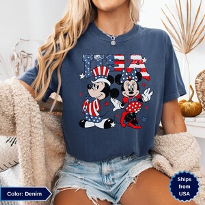 May include: Denim blue t-shirt featuring Mickey and Minnie Mouse in patriotic attire, with the word "USA" in a star-spangled design. Mickey wears a Uncle Sam hat, and Minnie has a bow. The shirt is a comfortable, casual style.