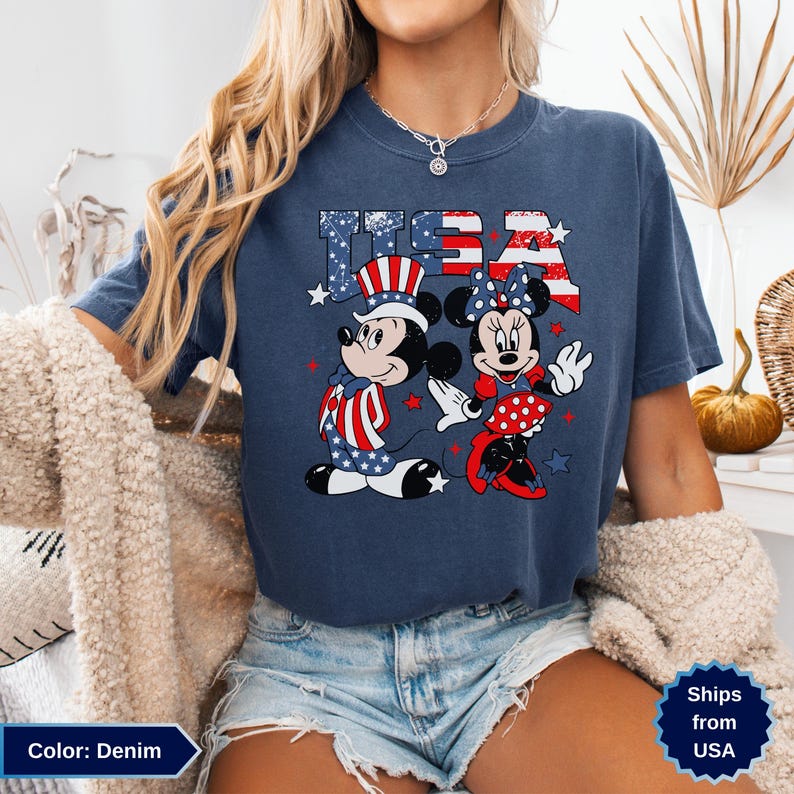 May include: Denim blue t-shirt featuring Mickey and Minnie Mouse in patriotic attire, with the word "USA" in a star-spangled design. Mickey wears a Uncle Sam hat, and Minnie has a bow. The shirt is a comfortable, casual style.