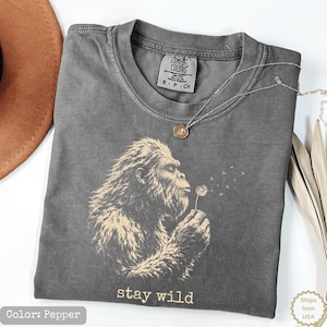 May include: A pepper-colored t-shirt featuring a graphic of a hairy creature blowing on a dandelion. The text "stay wild" is printed below the graphic. The shirt has a Comfort Colors label at the neck and is made of 100% cotton, size small.
