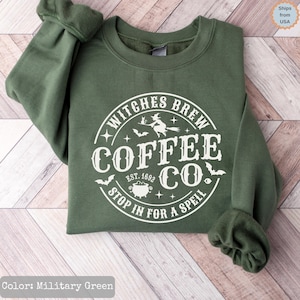 Könnte beinhalten: Ein militärgrüner Rundhals-Pullover mit einer weißen kreisförmigen Grafik und dem Text "WITCHES BREW COFFEE CO. EST. 1692 STOP IN FOR A SPELL". Das Design enthält Illustrationen einer Hexe auf einem Besen, eines Kessels, Fledermäusen und Sternen.