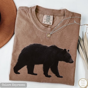 May include: An espresso-coloured t-shirt featuring a large, detailed graphic of a black bear walking across the centre. The shirt has a crew neckline with a Comfort Colors brand label inside, and a silver-toned chain necklace rests on the fabric.