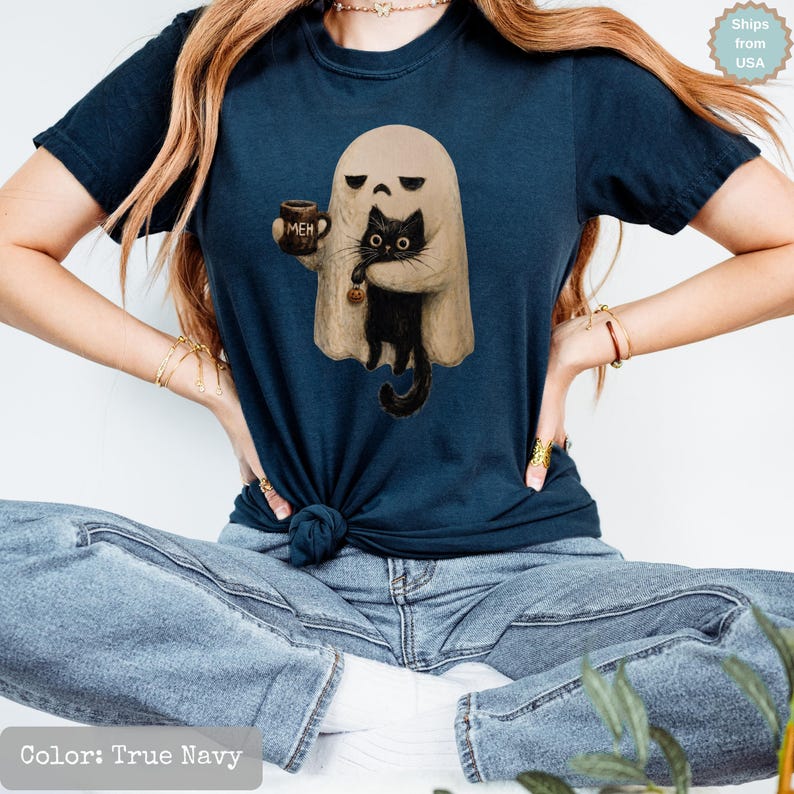 May include: A true navy short-sleeve t-shirt featuring a graphic of a beige ghost holding a black cat and a brown mug with the text MEH printed on it. The ghost has a bored expression, and the cat is looking forward with wide eyes.