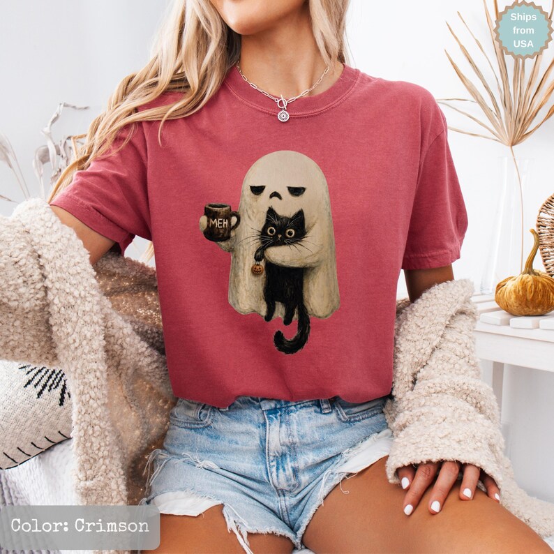 May include: A crimson t-shirt featuring a graphic of a beige ghost holding a brown mug with the text MEH and carrying a black cat. The ghost has a bored expression, and the cat has large eyes. The shirt has a crew neckline and short sleeves.