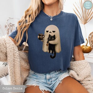 May include: A denim blue short-sleeve t-shirt featuring a graphic print of a beige ghost holding a brown mug with the text "MEH" and a black cat. The cat has large eyes and is holding a small orange pumpkin charm in its paws.