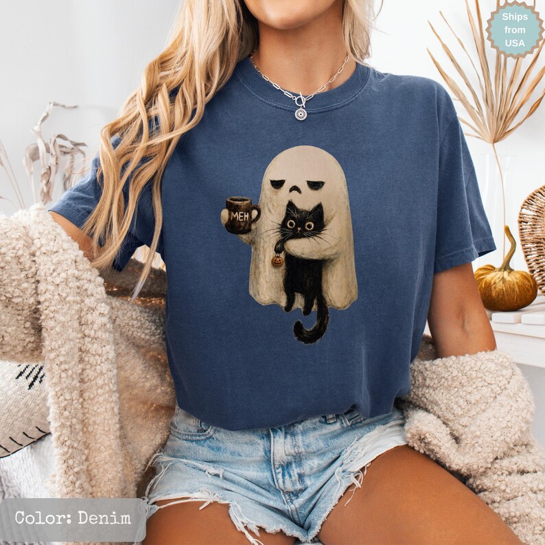 May include: A denim blue short-sleeve t-shirt featuring a graphic print of a beige ghost holding a brown mug with the text "MEH" and a black cat. The cat has large eyes and is holding a small orange pumpkin charm in its paws.