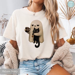May include: An ivory short-sleeve t-shirt featuring a graphic of a beige ghost holding a brown mug with the text MEH and carrying a black cat. The cat has large eyes and holds a small orange pumpkin. The shirt has a crew neckline.