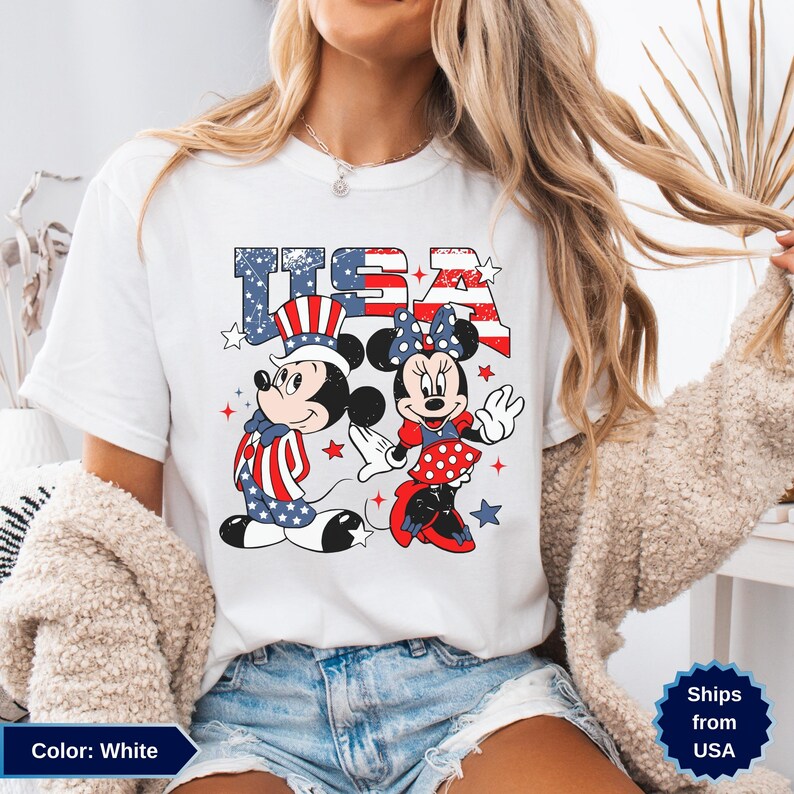 May include: White t-shirt featuring Mickey and Minnie Mouse in patriotic outfits with the text "USA" in red, white, and blue. Mickey wears a hat and suit with stars and stripes, while Minnie dons a polka-dot dress and bow.