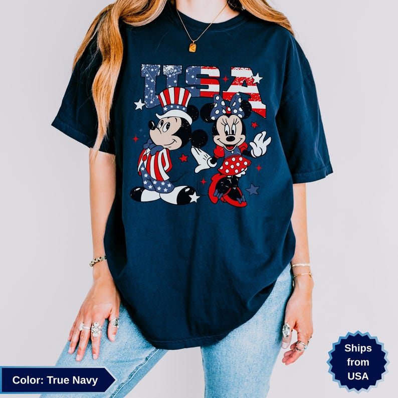 May include: True navy t-shirt featuring Mickey and Minnie Mouse in patriotic attire with the text "USA". Mickey wears a hat and outfit with red, white, and blue stars and stripes. Minnie wears a polka dot skirt and bow.