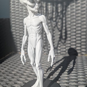 May include: A white, three-dimensional figurine of an extraterrestrial being with an elongated head, large dark almond-shaped eyes, and a slender, muscular physique. The figure stands upright on a circular black base, showcasing detailed anatomical features.