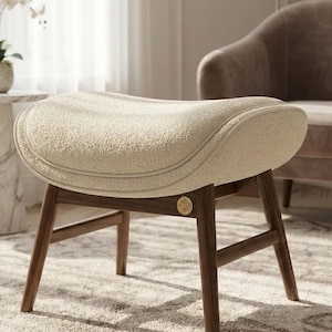 Puede incluir: Un otomano de tela texturizada color crema con una parte superior curva y acolchada. La base está construida en madera marrón oscuro con cuatro patas inclinadas y barras de soporte horizontales. Un pequeño emblema dorado está fijado al marco.