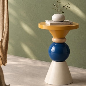 Puede incluir: Una mesa auxiliar con una superficie circular amarilla, un anillo beige, una esfera azul grande y una base cónica blanca. Sobre la mesa hay un libro titulado The Kinfolk Home y un pequeño jarrón blanco con una planta verde.