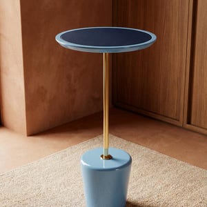 May include: A side table featuring a circular top with a dark blue textured surface and a light blue rim. The table is supported by a slender, vertical gold-toned metal rod connected to a solid, light blue, tapered cylindrical base with a glossy finish.