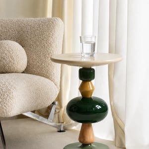 Puede incluir: Una mesa auxiliar con tablero redondo color crema y una base apilada que presenta un bloque rectangular verde oscuro, una pieza hexagonal amarillo mostaza, una forma redondeada verde oscuro, un cilindro de madera clara y una base circular verde salvia.