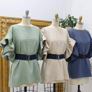 Op de afbeelding: Drie linnen tops in saliegroen, beige en marineblauw, elk met gerimpelde mouwen en een donkerblauwe riem met een rechthoekige gesp. De tops worden op mannequins getoond.