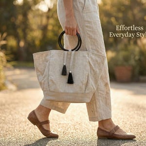 May include: A beige tote bag with black handles and black tassels is carried by a person wearing beige pants and brown shoes. The bag has a rectangular shape and is made of a textured fabric. The text "Effortless Everyday Style" is visible.