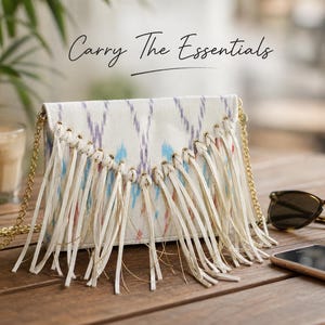 Könnte beinhalten: Eine weiße Clutch mit farbenfrohem Ikat-Muster und langen Fransen. Die Tasche hat einen goldfarbenen Kettenriemen und wird auf einem Holztisch mit Sonnenbrille und Smartphone präsentiert. Der Text "Carry The Essentials" steht über der Tasche.