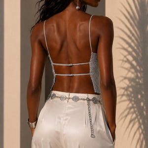 May include: A silver metallic backless camisole top with thin spaghetti straps and two horizontal back ties, paired with cream-colored satin trousers and a silver chain belt featuring circular coin-like charms and a dangling chain detail.