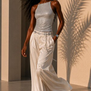 May include: A sleeveless silver sequined top with a high neckline paired with wide-leg cream satin trousers. The outfit includes a silver chain belt featuring circular medallions, long drop earrings with spherical pendants, and a wide silver cuff bracelet.