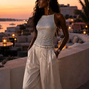 May include: A sleeveless silver sequined top with a high neckline paired with wide-leg cream satin trousers. A thin silver chain belt with circular accents rests on the waist, and a single long silver earring with a pearl drop is visible.