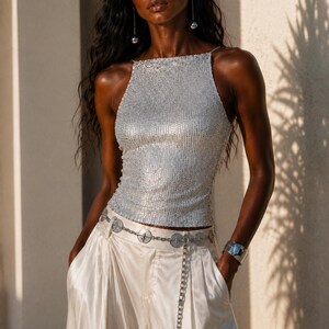 May include: A sleeveless silver sequined top with a high neckline paired with cream-colored satin wide-leg trousers. The outfit includes a silver chain belt featuring circular metal accents and a wide silver cuff bracelet on the left wrist.