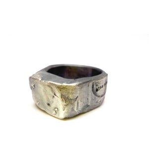 Könnte beinhalten: Ein silberfarbener Ring mit einer dicken, unregelmäßigen und texturierten Ringschiene. Die Oberfläche weist ein verwittertes, dunkles Metall-Finish mit sichtbaren Vertiefungen und einem kleinen, eingestanzten Mondsichel-Symbol auf.