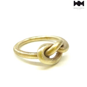 Könnte beinhalten: Ein goldener Knotenring mit glatter, matter Oberfläche. Der Ring besteht aus einem dicken Metallband, das zu einer Knotenform verdreht ist.