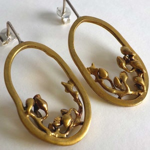 May include: A pair of gold-toned oval earrings with a botanical design. The earrings feature a branch-like structure with bud-shaped details within the oval frame. The earring posts are silver.
