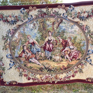 May include: A rectangular woven tapestry featuring a central oval scene of four figures in period clothing within a pastoral landscape with sheep and a waterfall. The border is cream with floral patterns and a deep burgundy outer frame.