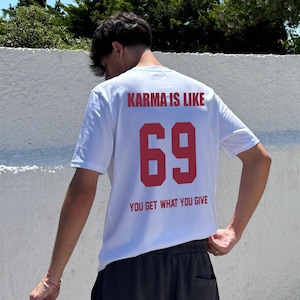 Può includere: Una t-shirt bianca a maniche corte con testo rosso che recita KARMA IS LIKE, il numero 69 in grande carattere rosso e il testo YOU GET WHAT YOU GIVE sottostante. La maglietta è abbinata a pantaloncini grigio scuro e un berretto nero in mano.