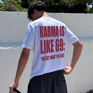 Può includere: Una t-shirt bianca a maniche corte con testo rosso sul retro che recita "KARMA IS LIKE 69: YOU GET, WHAT YOU GIVE". La maglietta è abbinata a pantaloncini grigio scuro e un cappellino nero tenuto in mano, mostrando uno stile casual.