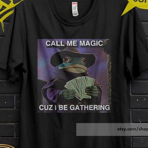 Può includere: T-shirt nero con una rana con un cappello che tiene dei soldi, con la scritta "CALL ME MAGIC CUZ I BE GATHERING". La maglietta ha anche la scritta "BUY 3 FREE" e "PREMIUM QUALITY".
